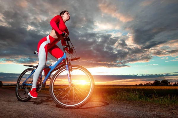 夕日を背景に自転車に赤いスポーツスーツの美しい女の子 健康的なライフスタイル スポーツトレーニング 心臓負荷の概念 スペースのコピー — ストック写真