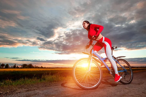 夕日を背景に自転車に赤いスポーツスーツの美しい女の子 健康的なライフスタイル スポーツトレーニング 心臓負荷の概念 スペースのコピー — ストック写真