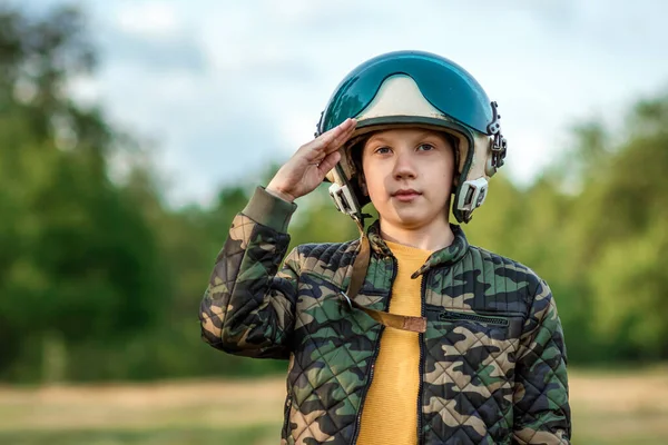 A boy in a pilot\'s helmet on a background of greenery salutes. Dream concept, choice of profession, military service. Copy space