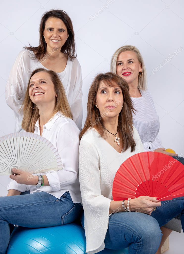 Group of 4 women, friends, middle-aged having fun in a photo session in a studio with white background, sitting on a ball and playing with a fan