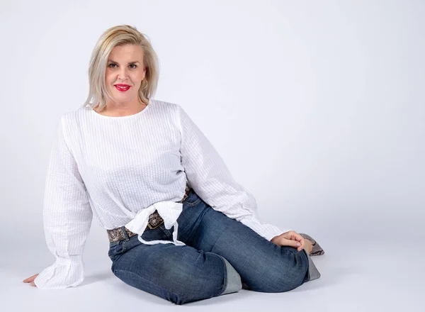 Photographic studio session for a 50 year old woman sitting on the floor and dressed in jeans and white shirt.