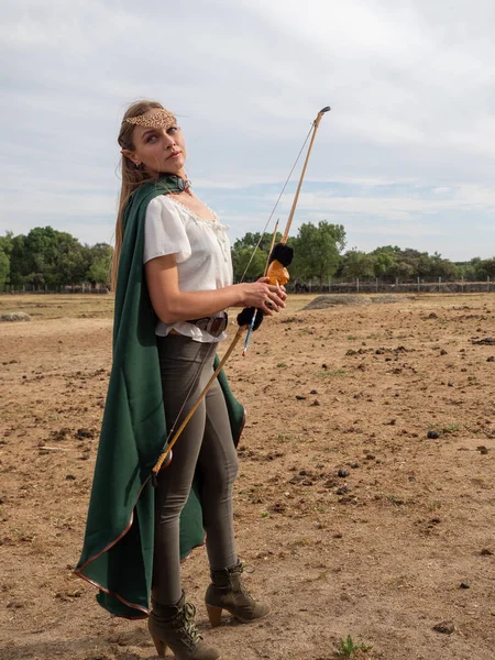 Blonde girl with elf ears poses in the field with a bow and a green cape.