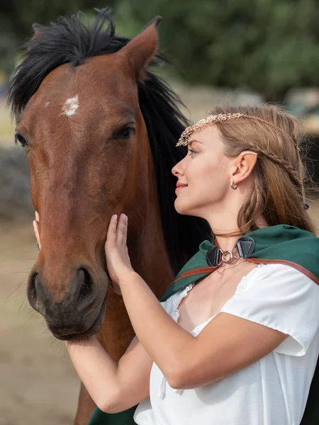 Blonde girl with blue eyes and makeup with elf in the field with a brown horse and a green cape.
