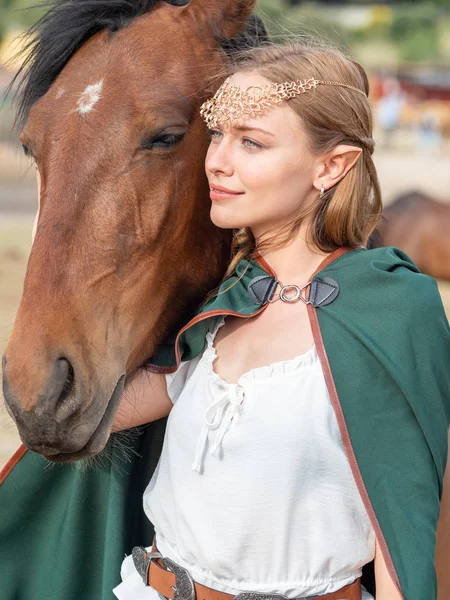 Blonde girl with blue eyes and makeup with elf in the field with a brown horse and a green cape.