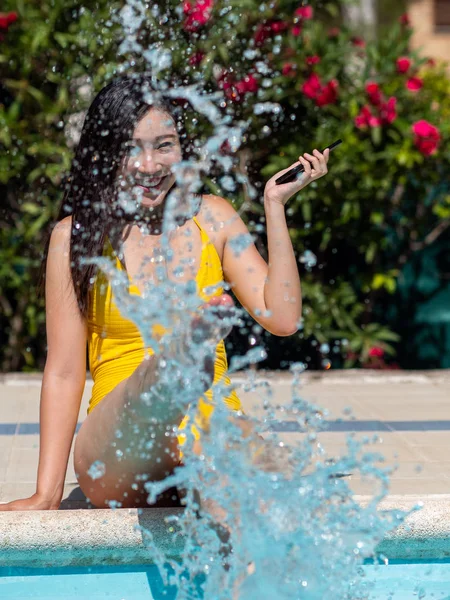 Aziatisch meisje in gele bikini neemt de zon en communiceert met haar mobiele telefoon naast een zwembad. — Stockfoto