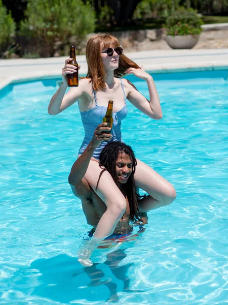 異人種間のカップルがプールで遊ぶ。彼女は彼の肩の上にあり、彼らはビールを飲む — ストック写真