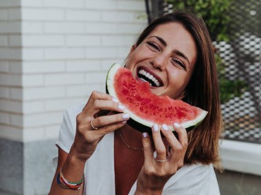 Beyaz kadın evinin bahçesinde karpuz dilimleri yiyor ve yerde oturuyor.