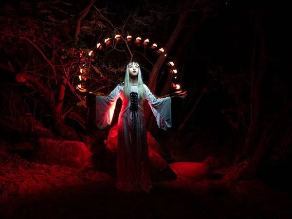 Una mujer sentada en una roca en el bosque y vestida con una túnica, arroja luces rojas de sus manos . — Foto de Stock