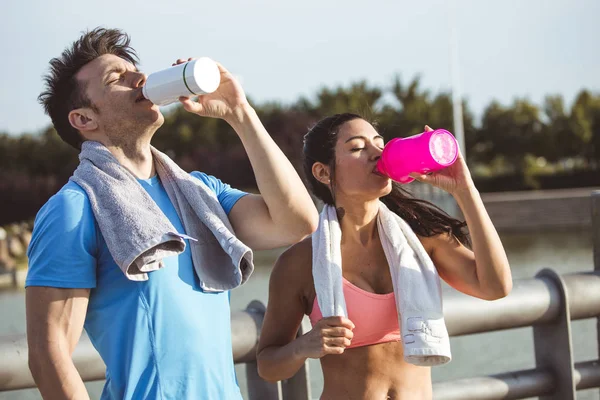 Pareja interracial, latina y caucásica beben de una botella de agua con una toalla alrededor de su cuello después de los deportes al aire libre Fotos De Stock Sin Royalties Gratis