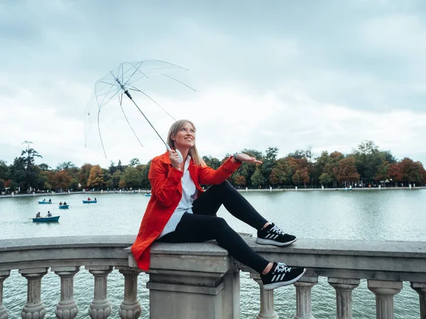 Ragazza caucasica con gabardine rosse e ombrello trasparente seduta accanto al lago Retiro Park a Madrid, Spagna . — Foto Stock