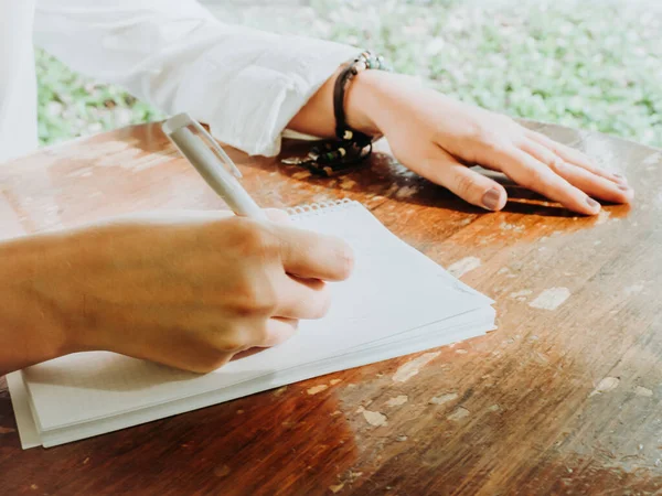 Blank meisje neemt notities in een notitieboekje op een houten tafel naast een raam. — Stockfoto