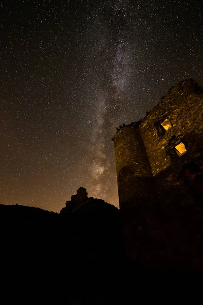 Dlouhá expozice noční fotografie s Mléčnou dráhou v pozadí, mezi horami a vedle budovy s osvětlenými okny. — Stock fotografie