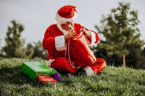 Foto de estoque vertical de Papai Noel sem barba agachado no chão organizando presentes com sua bolsa vermelha. Tempo de Natal — Fotografia de Stock