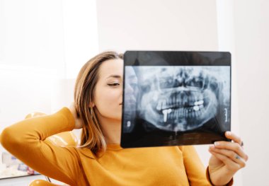 Diş kliniğinde sandalyede otururken gülümseyen ve diş röntgeni çeken genç bir kadın.