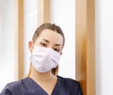 Maskeli ve önlüklü genç bir kadın modern hastanede çalışırken kameraya bakıyor.