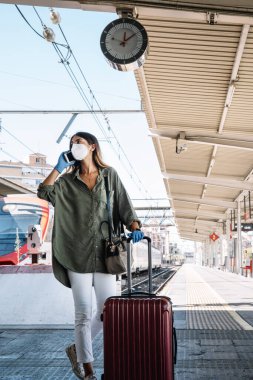 Lastik eldivenli genç bir kadın valiz çekiyor ve salgın sırasında tren istasyonunda yürürken cep telefonu kullanıyor.