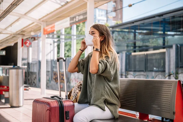 Genç bayan gezgin bavulun yanında bankta oturuyor ve salgın sırasında treni beklerken tıbbi maske takıyor.