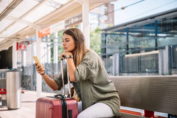 Genç bayan turist yaz tatilinde tren istasyonunda bankta otururken bavula yaslanıyor ve akıllı telefona bakıyor.