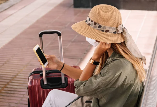 Side view of unrecognizable woman in hat and mask sitting on bench near suitcase and browsing smartphone on railroad station during pandemic