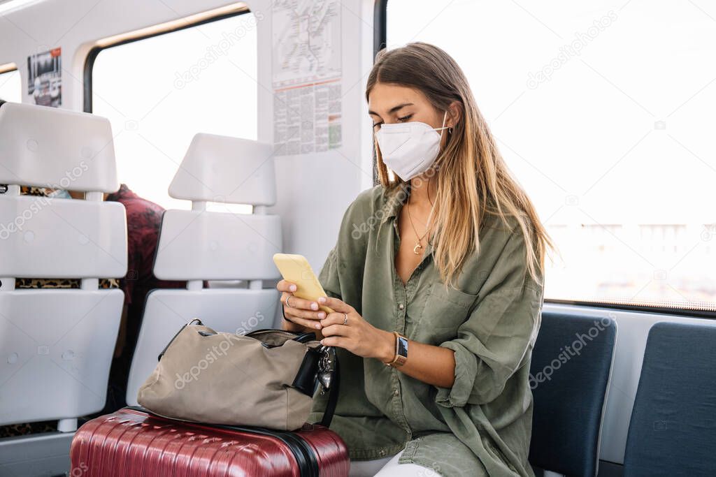 Young female traveler in medical mask and with suitcase looking to the smartphone while riding contemporary train during pandemic