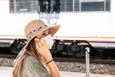 Şapkalı ve maskeli kadın gezgin, gözünü kaçırıyor ve tren istasyonunda salgın sırasında trenle ilgili akıllı telefondan konuşuyor.