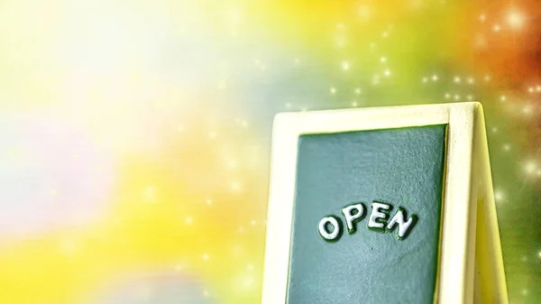 Close up of a green with yellow "OPEN" chalkboard sign leaning against sparkle ,bright with colorful background. — Stock Photo, Image