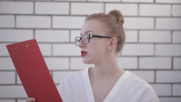 Porträt einer Bürofrau mit Brille, weißem Hemd und roten Lippen, die einen Ordner mit Papieren hält und Dokumente auf weißem Hintergrund liest — Stockvideo