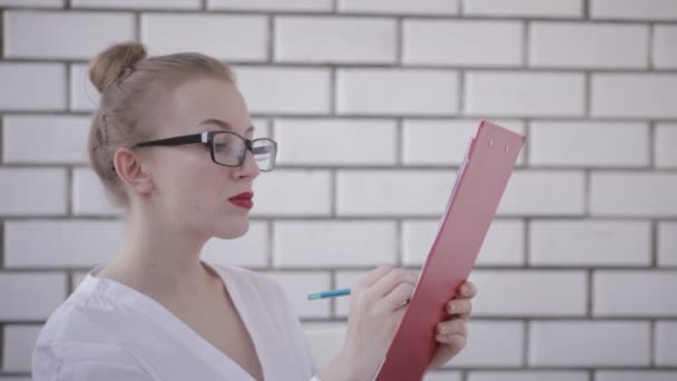 Портрет офисной женщины в очках с белой рубашкой и красными губами, держащей папку с бумагами и пишущей документы изолированы на белом фоне — стоковое видео