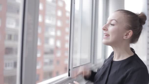 Усміхнена дівчина стоїть і махає біля вікна — стокове відео