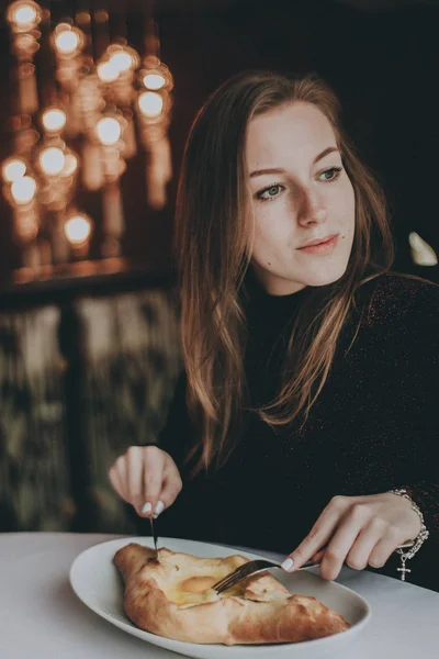 Güzel bir kız hafif bir restoranda oturur ve bir bıçak ve çatal ile bir sandviç yiyor. Kadın kahvaltı café'de yaşıyor. Sandviç bir bıçak ile kesmek.