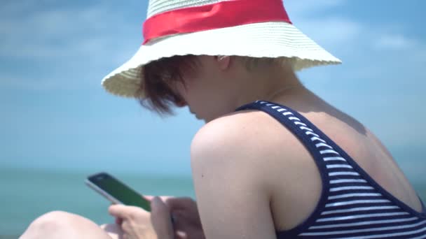 Joven hermosa mujer tipos de texto en la aplicación de mensajero usando teléfono inteligente con sombrero retro y traje de baño con rayas azules y blancas en la playa de verano — Vídeos de Stock