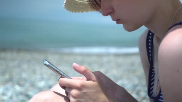 Mujer joven y bonita usando teléfono inteligente junto a la playa del mar. Chica en sombrero retro y traje de baño con rayas azules y blancas. Concepto de tecnología y descanso . — Vídeos de Stock