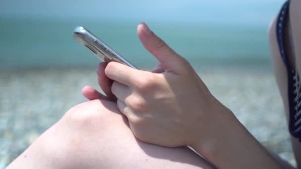 年轻女子手在海边模糊背景使用智能手机的特写镜头。休息和技术。网上冲浪 — 图库视频影像