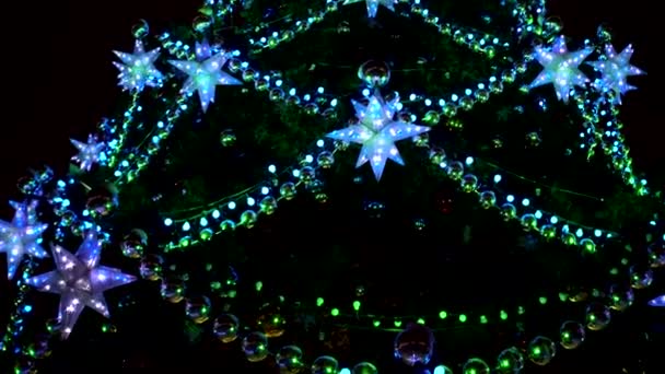 Cidade magnífica Árvore de Natal em quadrado brilhando com luzes verdes, azuis e amarelas de guirlandas, bolas e estrelas — Vídeo de Stock