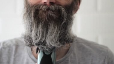 Yarı gri sakallı orta yaşlı bir adam tıraş makinesiyle sakal kesmeye çalışıyor.
