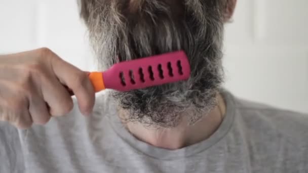 Homem de meia-idade com barba cinza penteando barba longa no banheiro por pente rosa — Vídeo de Stock