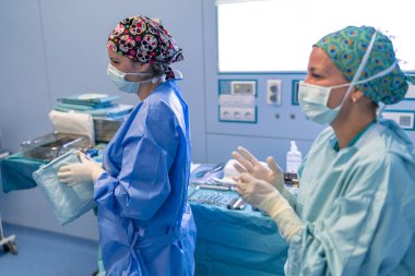 Cerrah yardımcı olmak bekleyen ameliyathane yardımcıları, steril forma giy. Sol profil maskesi altında gülümseyen girdiler. Sen-ebilmek görmek onları arkasında cerrahi tablo.