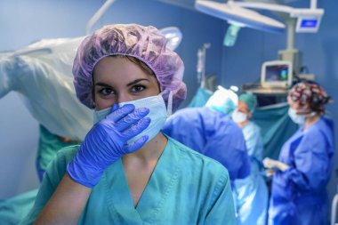 Cerrahi asistanı kameraya eliyle maske üzerinde arka planda çalışma cerrahi ekibi ile arıyorum. Mor şapka giyiyor.