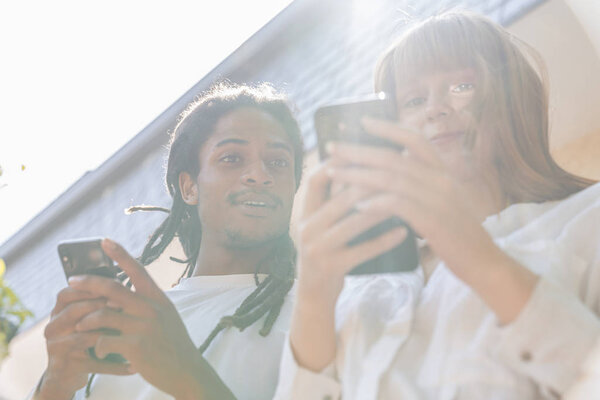 Afro boy and redheaded Caucasian girl chatting on the mobile phone on the terrace, him looking at her mobile. Sunny day. Image from below