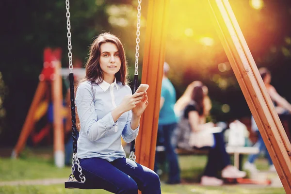 Girl student teenager swinging on a swing and using phone for communication looks in frame.