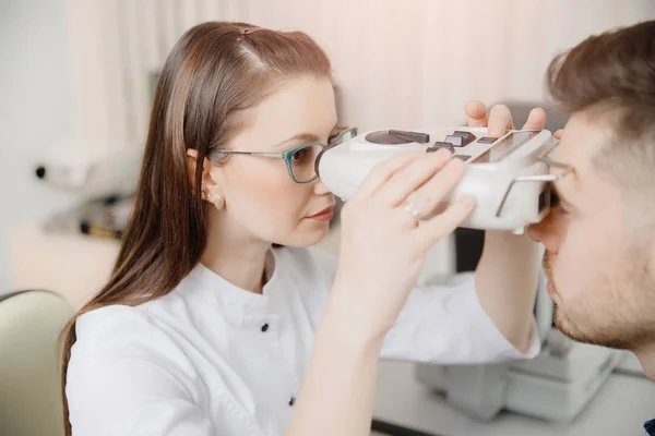 Médico de menina atraente oftalmologista verifica a visão do jovem com corte de cabelo na moda e barba em equipamento especial — Fotografia de Stock