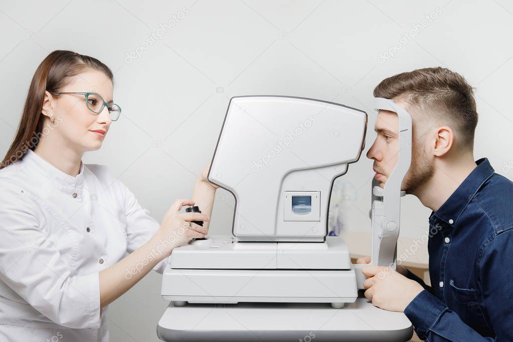 Attractive dark-haired young woman doctor measures eye pressure on ophthalmic equipment to fashionable guy in blue shirt