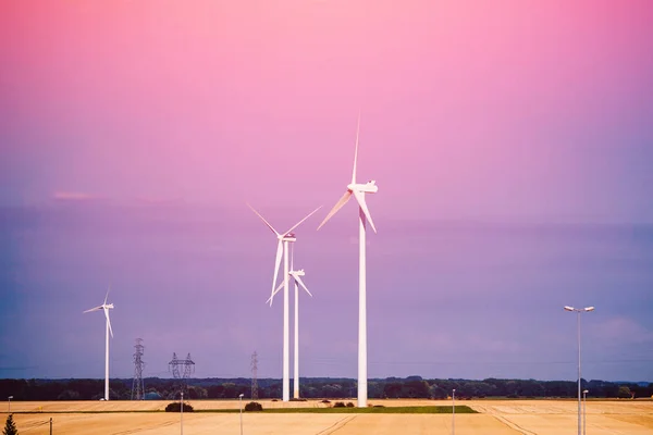 タービンブレードを備えた複数の風力発電機は、日没時に黄色いフィールドの夕方に立っています — ストック写真