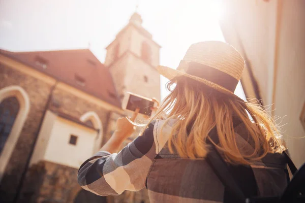 Молодая женщина туристка в соломенной шляпе и с рюкзаком использует телефон в качестве камеры, фотографируя здания — стоковое фото