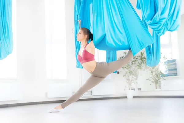 Ung flicka gymnast i sportkläder som sträcker sig på blå hängmatta i vit studio. Begreppet flygande yoga — Stockfoto