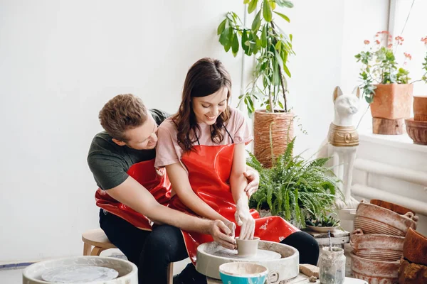 Tenero abbraccio di coppia romantica e giovane in grembiuli rossi al volante ceramista fare piatti in ceramica. Concetto famiglia — Foto Stock