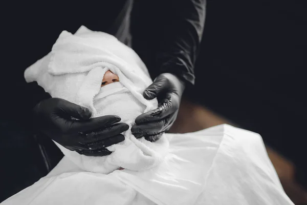 Man zit in kappersstoel en stoom zijn gezicht met hete handdoek voor koninklijke baard scheermes — Stockfoto