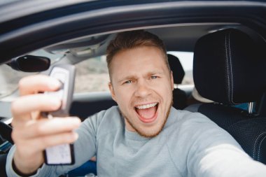 Yeni araba galerisinde oturan yakışıklı genç şoför gülümsüyor ve selfie çekiyor. Yaşam tarzı seyahatini kavra