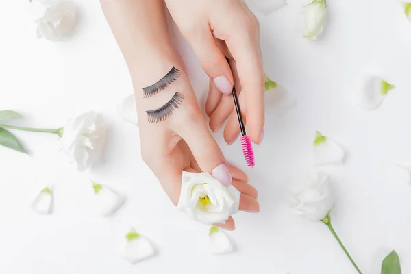 Falsas pestañas en la mano mujer maestro extensión flores blancas en el fondo. Concepto belleza moda —  Fotos de Stock