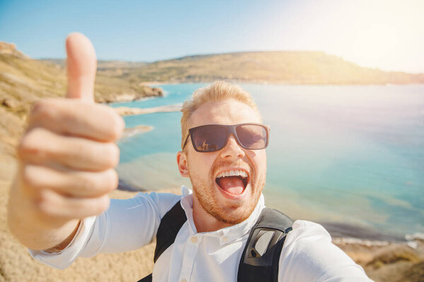Мужчина делает селфи фото на фоне синего моря, песчаного пляжа в солнечных очках и с рюкзаком. Концепция путешествия
